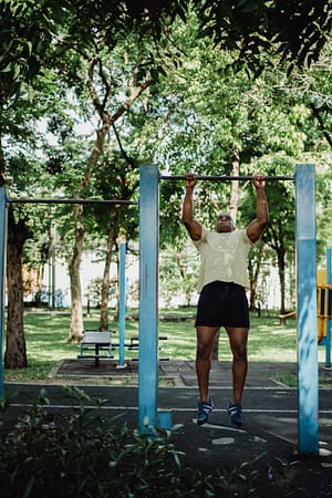 Services African American man doing pull-ups on outdoor exercise equipment in a sunny park setting.